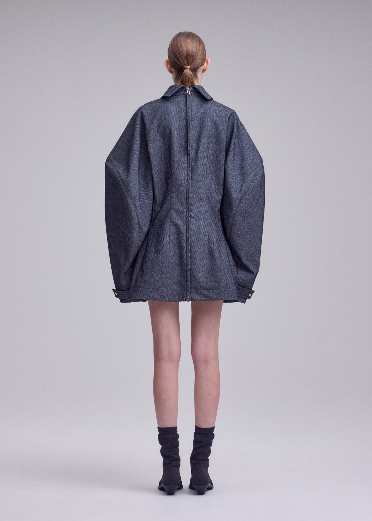 A person stands facing away, wearing the ISSEY MIYAKE REFLECTION oversized gray jacket with extra-wide sleeves and a short hem, layered over a silver high-neck tunic, paired with black ankle boots and socks. The background is plain and light grey. view 5 of 8