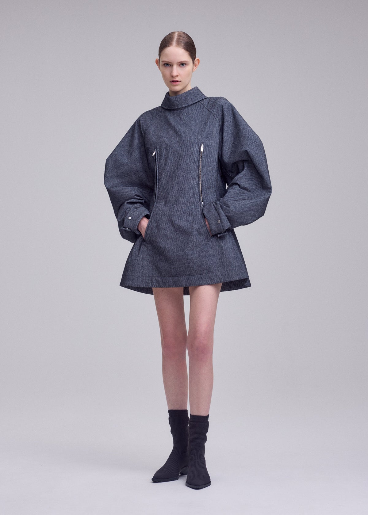 A model stands against a plain background, wearing the ISSEY MIYAKE REFLECTION oversized grey jacket featuring wide sleeves and front zippers, paired with black ankle boots. Beneath the jacket, she wears a silver high-neck tunic from the same collection, crafted from reflective tunic fabric that subtly catches the light. The model’s hair is pulled back, and her hands rest in the jacket pockets. view 6 of 8