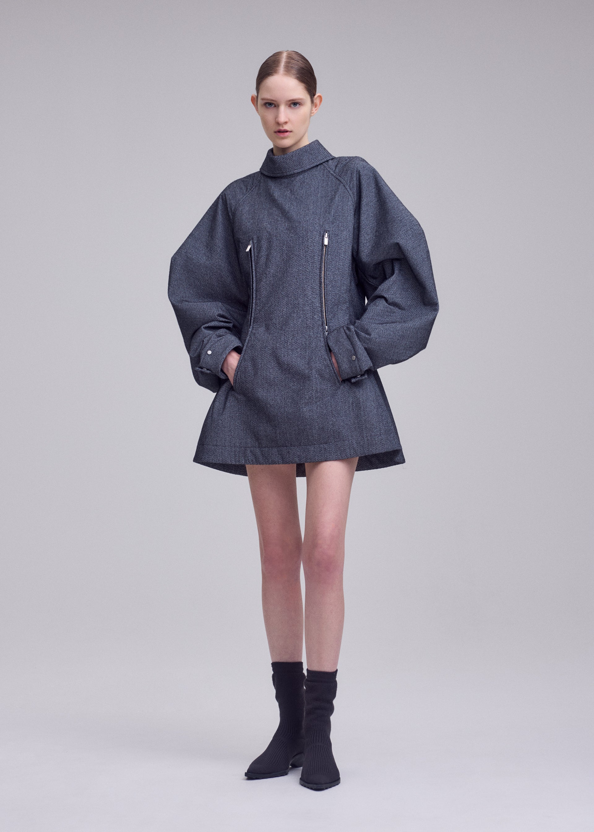 Zoomed view A model stands against a plain background, wearing the ISSEY MIYAKE REFLECTION oversized grey jacket featuring wide sleeves and front zippers, paired with black ankle boots. Beneath the jacket, she wears a silver high-neck tunic from the same collection, crafted from reflective tunic fabric that subtly catches the light. The model’s hair is pulled back, and her hands rest in the jacket pockets.