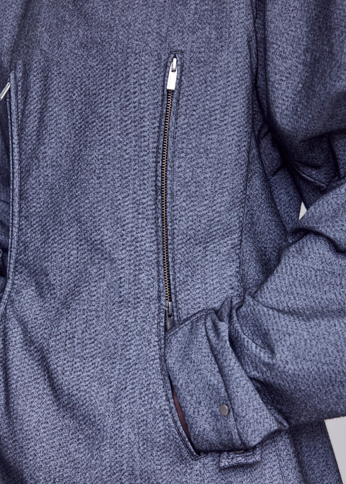 Close-up of a person wearing the ISSEY MIYAKE REFLECTION jacket—a textured gray piece with a diagonal zipper and right hand tucked into a side pocket—featuring subtle reflector fabric that enhances both style and visibility. view 7 of 8