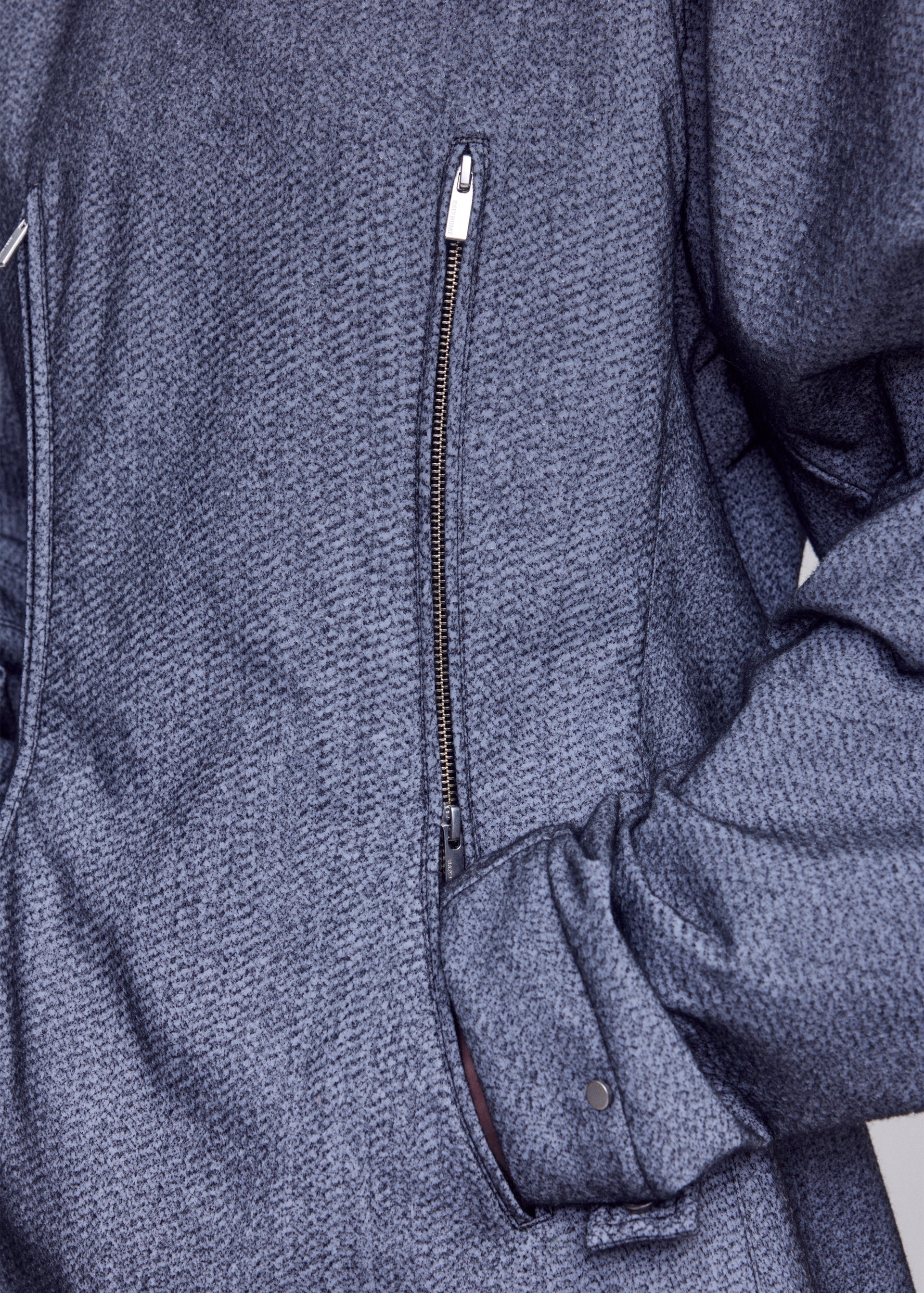 Zoomed view Close-up of a person wearing the ISSEY MIYAKE REFLECTION jacket—a textured gray piece with a diagonal zipper and right hand tucked into a side pocket—featuring subtle reflector fabric that enhances both style and visibility.