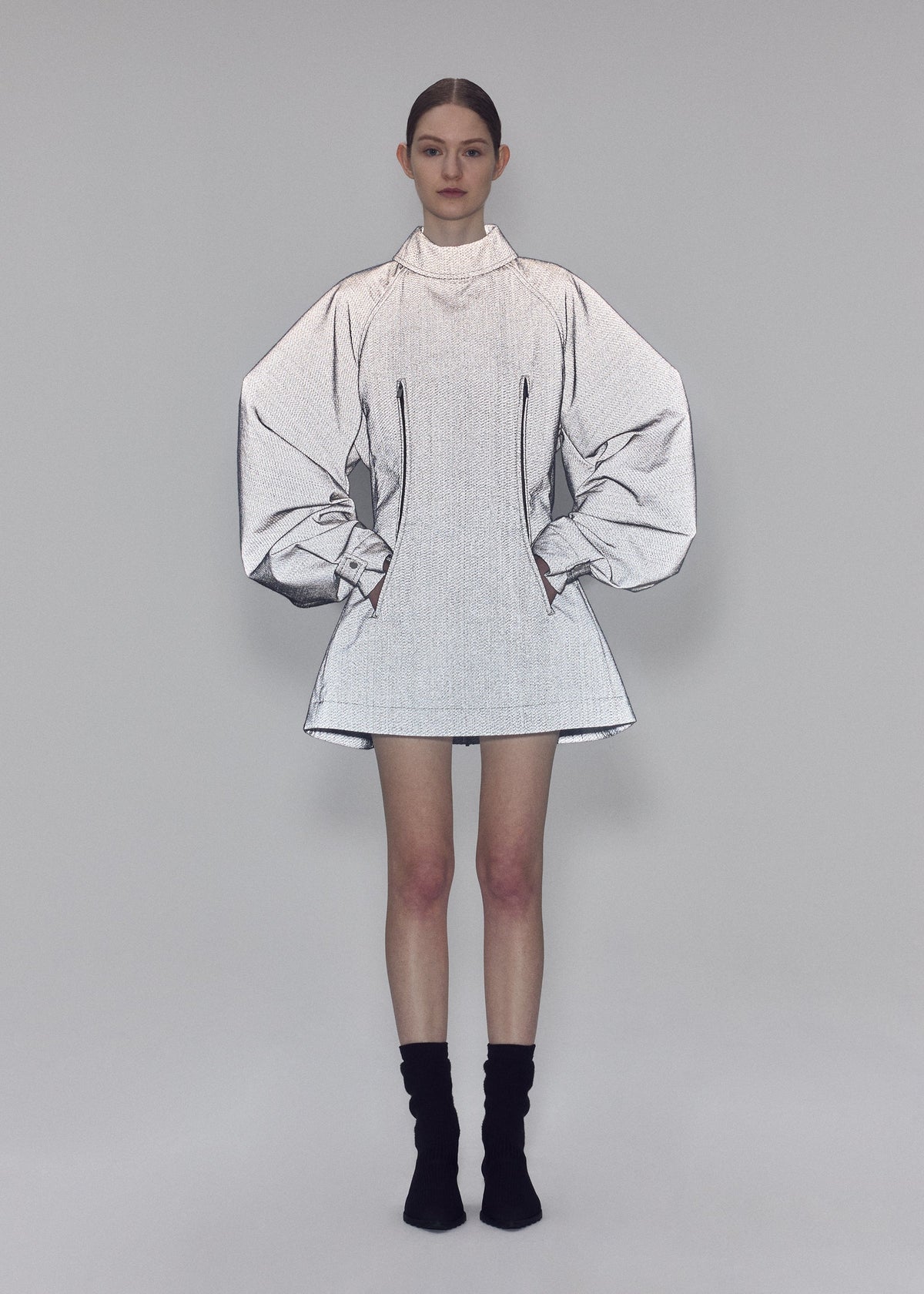 A woman stands against a gray background wearing the REFLECTION silver high-neck tunic by ISSEY MIYAKE, featuring exaggerated, sketch-like sleeves and a bodice crafted from reflector fabric. She pairs it with black ankle socks and black shoes, her hands in her pockets and her hair neatly tied back. view 8 of 8