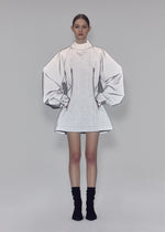 A woman stands against a gray background wearing the REFLECTION silver high-neck tunic by ISSEY MIYAKE, featuring exaggerated, sketch-like sleeves and a bodice crafted from reflector fabric. She pairs it with black ankle socks and black shoes, her hands in her pockets and her hair neatly tied back.