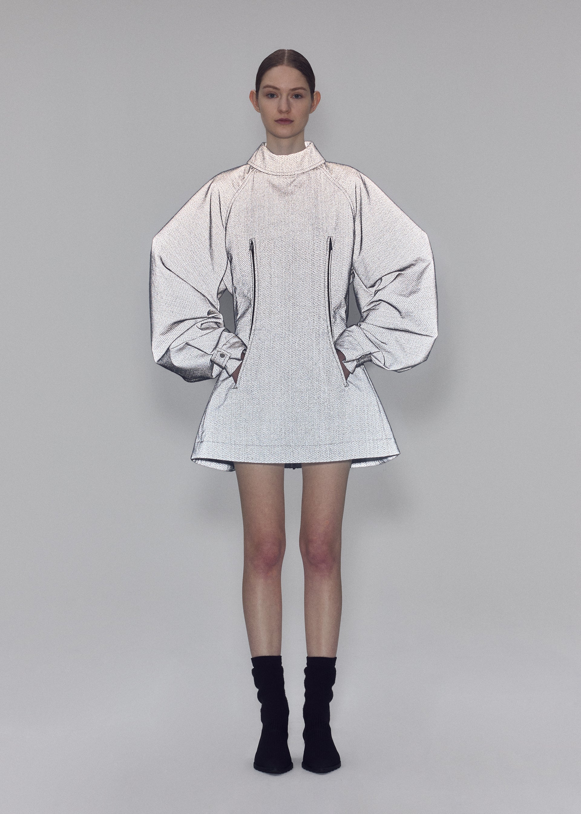 Zoomed view A woman stands against a gray background wearing the REFLECTION silver high-neck tunic by ISSEY MIYAKE, featuring exaggerated, sketch-like sleeves and a bodice crafted from reflector fabric. She pairs it with black ankle socks and black shoes, her hands in her pockets and her hair neatly tied back.