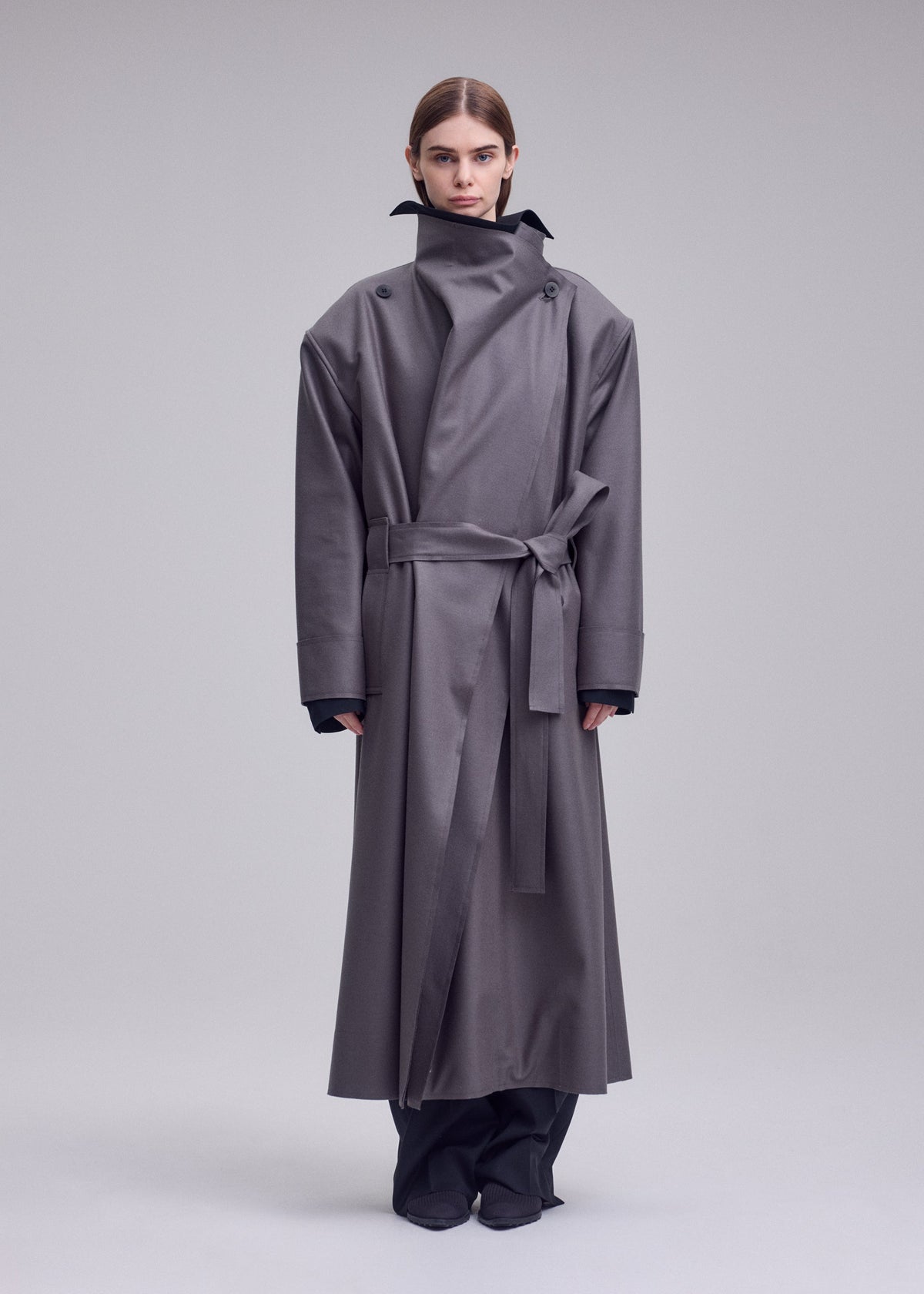 A person stands facing forward, wearing the SHELL coat by ISSEY MIYAKE. This long, oversized gray trench features wide shoulders, a high collar, and a belt cinched at the waist—crafted in ISSEY MIYAKE’s signature style. The background is plain and light gray. view 5 of 7