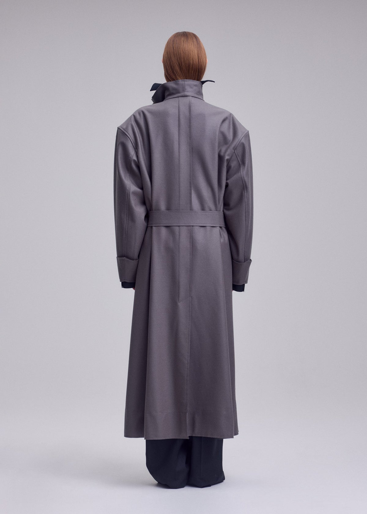 A person stands facing away, wearing ISSEY MIYAKE’s SHELL—a long, oversized gray coat with a belted waist and high collar, crafted from soft jersey material that exemplifies the brand’s distinctive style. Their straight, reddish-brown hair is visible above the collar. The background is plain and light gray. view 6 of 7