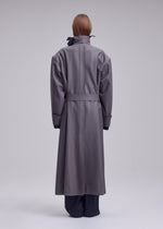 A person stands facing away, wearing ISSEY MIYAKE’s SHELL—a long, oversized gray coat with a belted waist and high collar, crafted from soft jersey material that exemplifies the brand’s distinctive style. Their straight, reddish-brown hair is visible above the collar. The background is plain and light gray.