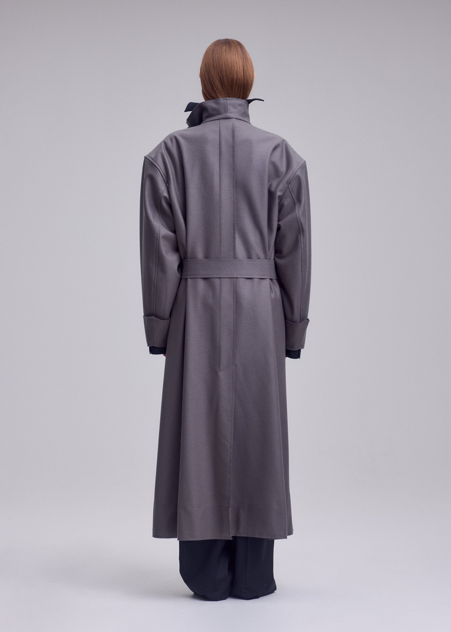 Zoomed view A person stands facing away, wearing ISSEY MIYAKE’s SHELL—a long, oversized gray coat with a belted waist and high collar, crafted from soft jersey material that exemplifies the brand’s distinctive style. Their straight, reddish-brown hair is visible above the collar. The background is plain and light gray.