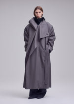 A person stands against a plain background wearing the SHELL, a long, oversized gray coat by ISSEY MIYAKE featuring a wide collar. They pair it with a soft black jersey turtleneck and loose black pants. Their hands are in the coat pockets and their expression is neutral.