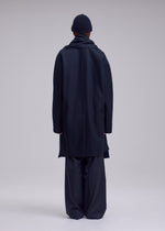 A person stands facing away, wearing the ISSEY MIYAKE MANNER wool jersey coat with wide-leg pants, scarf, and beanie against a plain, light background.