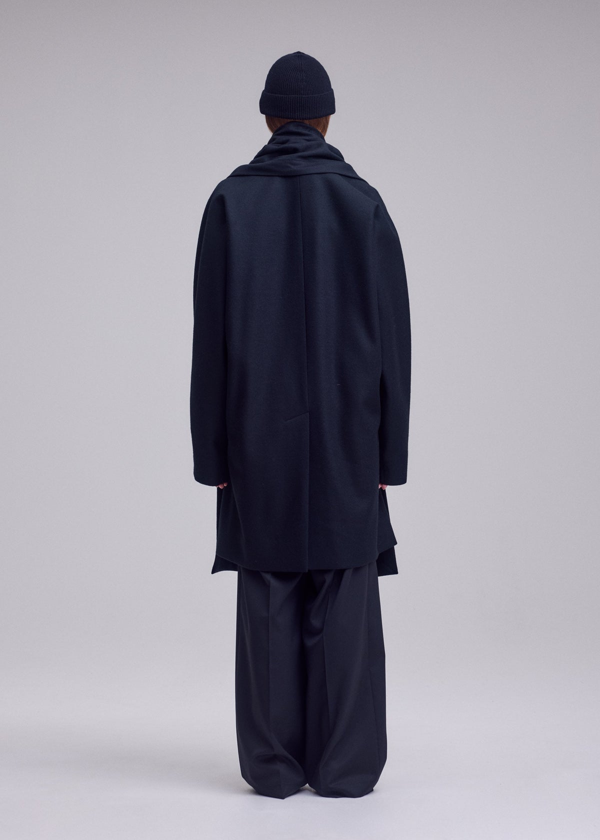 Zoomed view A person stands facing away, wearing the ISSEY MIYAKE MANNER wool jersey coat with wide-leg pants, scarf, and beanie against a plain, light background.