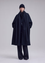 A person stands against a plain background wearing the ISSEY MIYAKE MANNER black recycled fabric coat with a beanie, scarf, pants, and shoes, hands in pockets and a neutral expression.