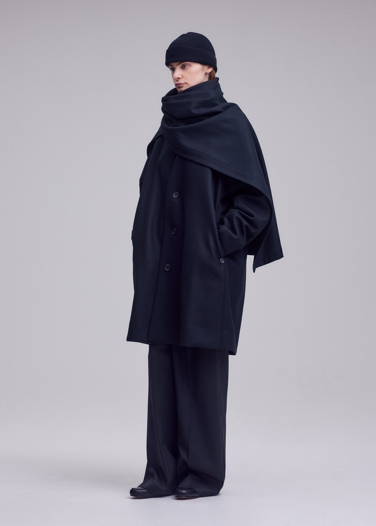A person wearing the ISSEY MIYAKE MANNER oversized lamb’s wool coat, wide-leg trousers, matching scarf, and a black knit beanie stands against a plain light gray background. view 8 of 8