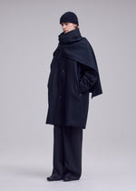A person wearing the ISSEY MIYAKE MANNER oversized lamb’s wool coat, wide-leg trousers, matching scarf, and a black knit beanie stands against a plain light gray background.