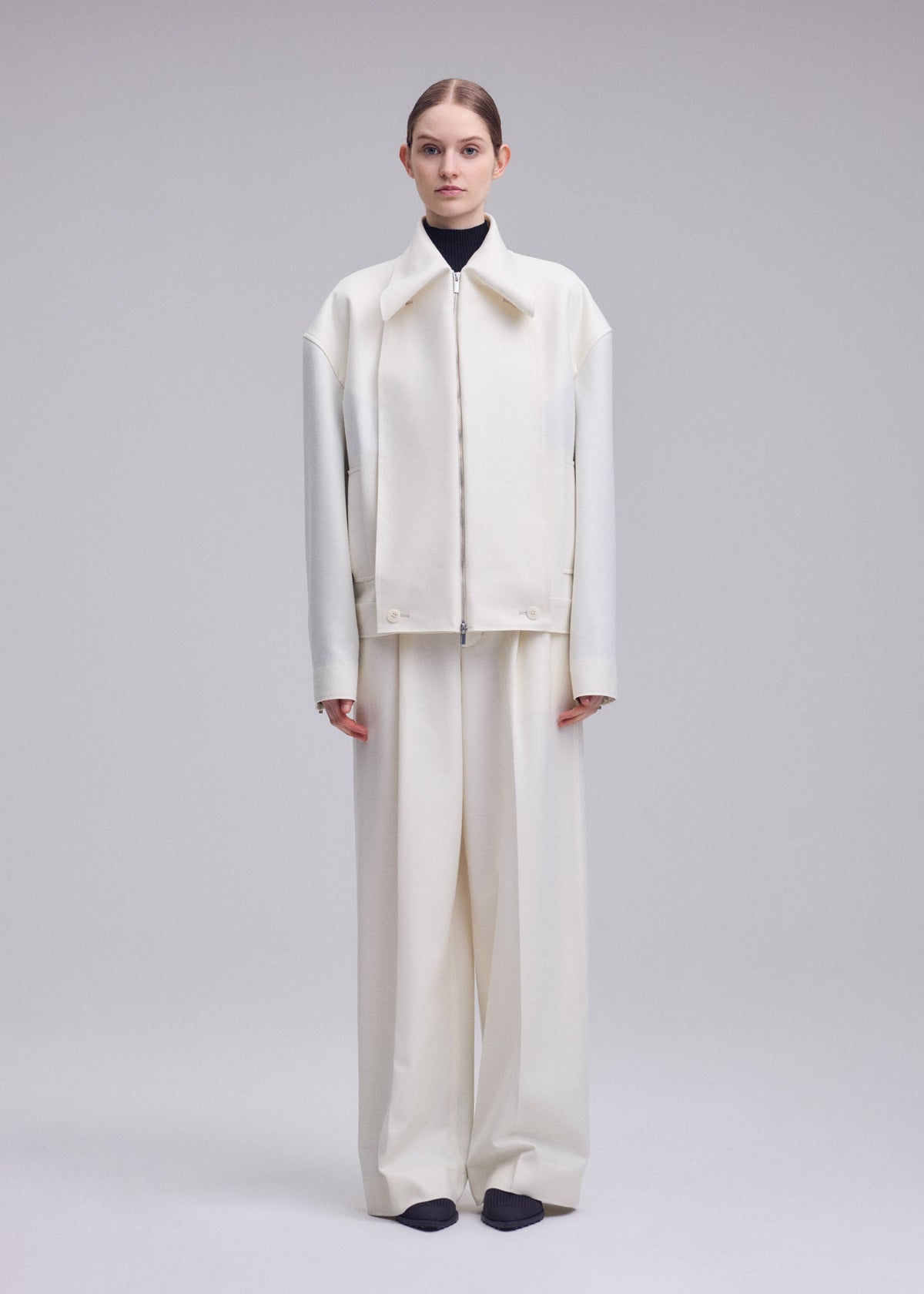 A person stands facing forward, wearing an oversized off-white SHELL jacket and wide-leg pants in luxurious wool jersey by ISSEY MIYAKE over a dark turtleneck, with black shoes, against a plain light gray background—a refined look from the Autumn Winter 2026/27 Collection. view 5 of 8