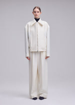 A person stands facing forward, wearing an oversized off-white SHELL jacket and wide-leg pants in luxurious wool jersey by ISSEY MIYAKE over a dark turtleneck, with black shoes, against a plain light gray background—a refined look from the Autumn Winter 2026/27 Collection.