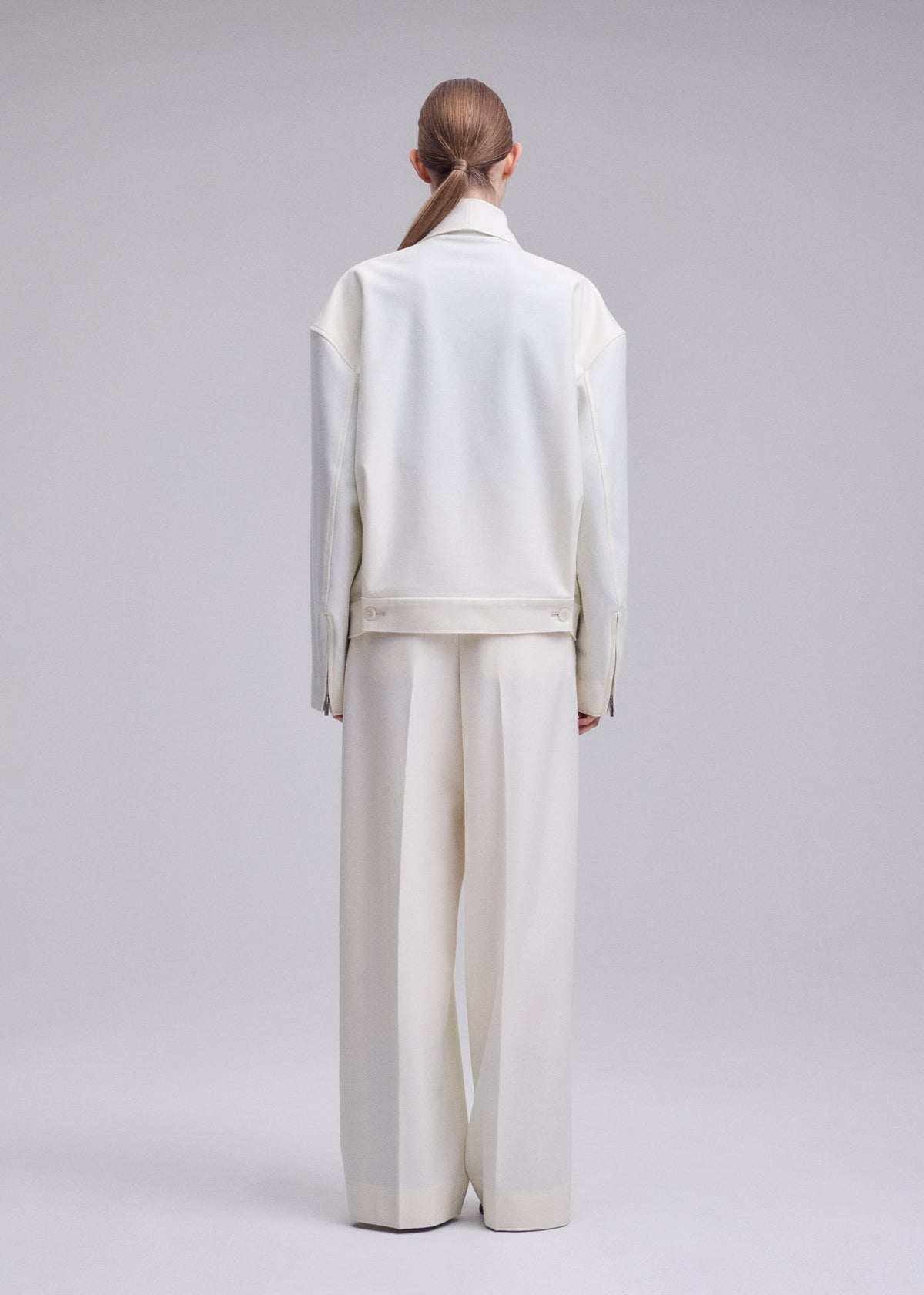 A person with light brown hair in a low ponytail is standing with their back to the camera, wearing an oversized ISSEY MIYAKE SHELL jacket from the Autumn Winter 2026/27 Collection and wide-leg white pants crafted from luxurious wool jersey against a light gray backdrop. view 6 of 8