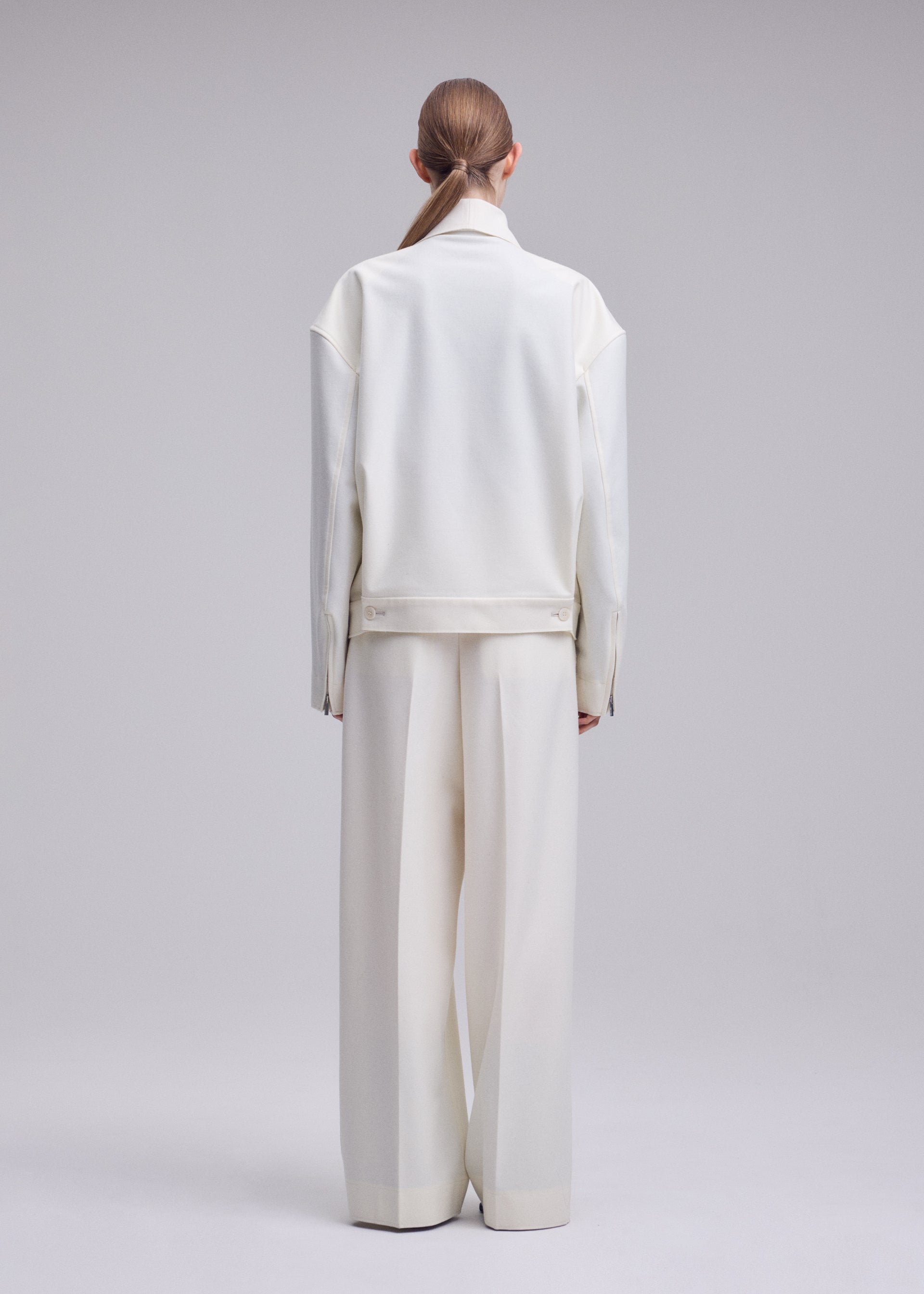 Zoomed view A person with light brown hair in a low ponytail is standing with their back to the camera, wearing an oversized ISSEY MIYAKE SHELL jacket from the Autumn Winter 2026/27 Collection and wide-leg white pants crafted from luxurious wool jersey against a light gray backdrop.