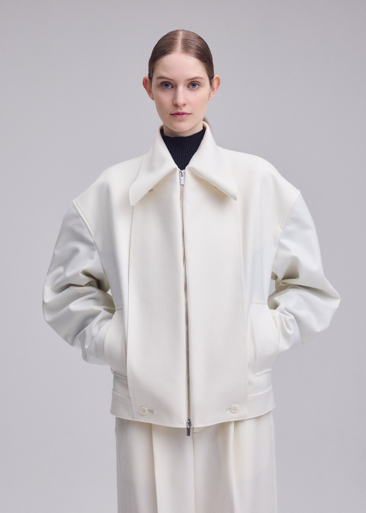 A person stands facing forward, wearing the white oversized SHELL jacket by ISSEY MIYAKE, crafted from luxurious wool jersey and paired with matching wide-leg pants. With hands in pockets and hair pulled back against a plain light background, this look exemplifies elegance from the Autumn Winter 2026/27 Collection. view 7 of 8