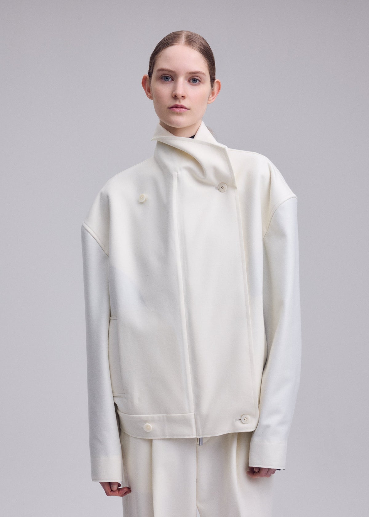 A person with light skin and brown hair, styled back, wears an oversized off-white SHELL jacket by ISSEY MIYAKE from the Autumn Winter 2026/27 Collection, crafted from luxurious wool jersey with a high collar and matching pants, standing against a plain light gray background. view 8 of 8
