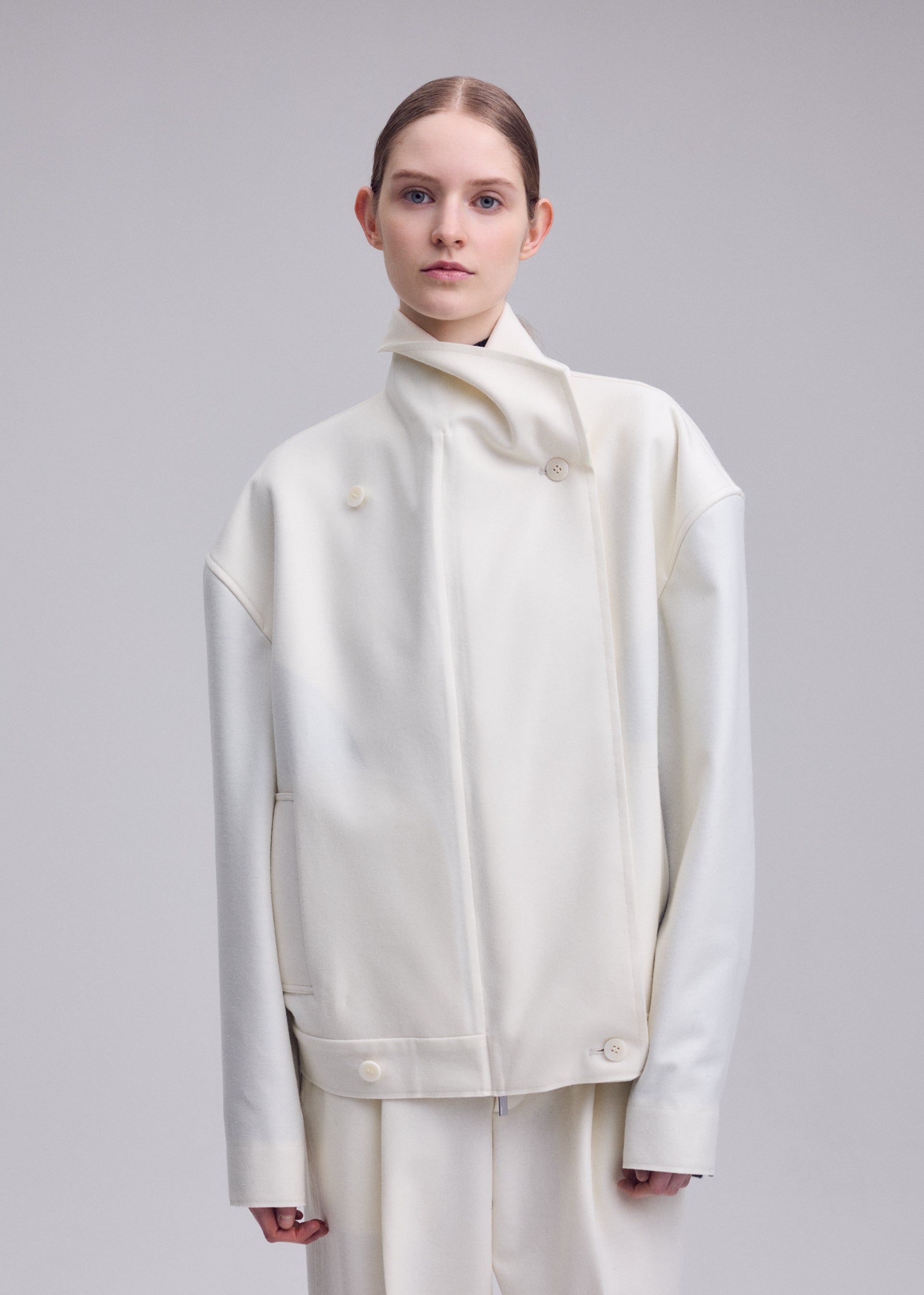 Zoomed view A person with light skin and brown hair, styled back, wears an oversized off-white SHELL jacket by ISSEY MIYAKE from the Autumn Winter 2026/27 Collection, crafted from luxurious wool jersey with a high collar and matching pants, standing against a plain light gray background.