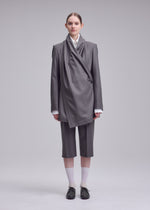 A person stands against a plain background wearing the ISSEY MIYAKE MANNER gray draped blazer and knee-length shorts, paired with a white shirt, black shoes, white socks; hair tied back with a neutral expression.