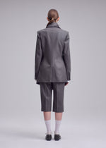 Person stands against a plain backdrop, facing away, wearing ISSEY MIYAKE’s MANNER—a long grey blazer and matching knee-length shorts—paired with white socks, dark shoes, and hair neatly tied back.