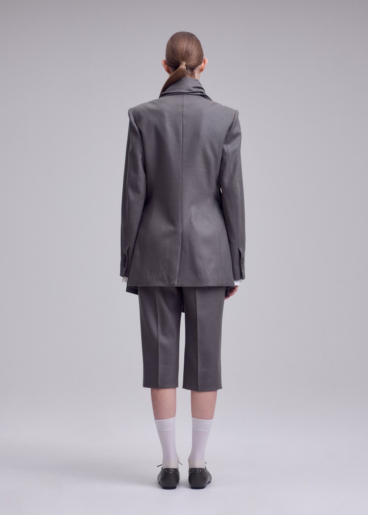 Zoomed view Person stands against a plain backdrop, facing away, wearing ISSEY MIYAKE’s MANNER—a long grey blazer and matching knee-length shorts—paired with white socks, dark shoes, and hair neatly tied back.