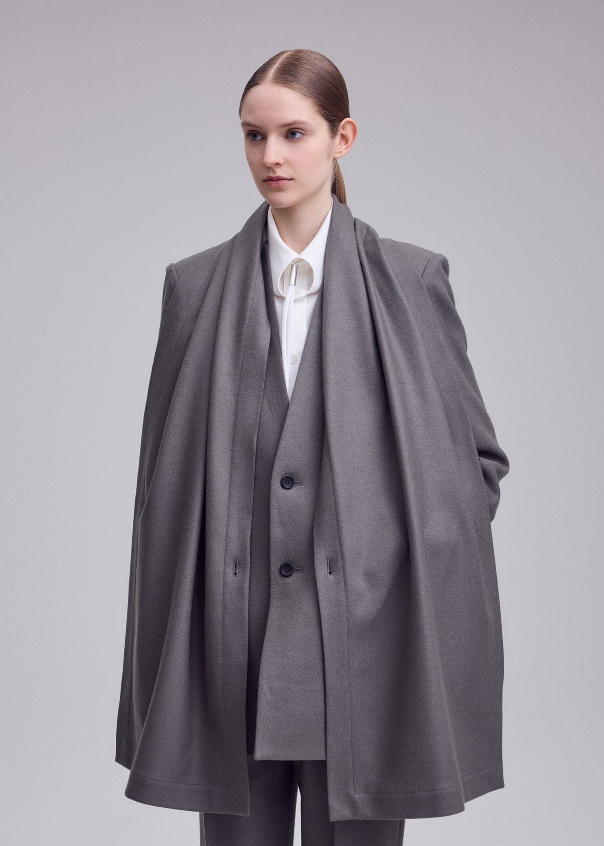 A woman in the ISSEY MIYAKE MANNER grey lamb’s wool jacket with a matching long scarf stands against a plain background, embodying sustainable fashion with her calm, neutral expression. view 7 of 8