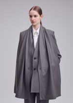 A woman in the ISSEY MIYAKE MANNER grey lamb’s wool jacket with a matching long scarf stands against a plain background, embodying sustainable fashion with her calm, neutral expression.