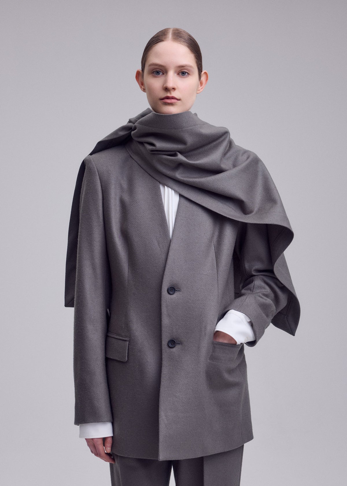 A person stands against a plain background wearing the MANNER grey oversized blazer and matching pants by ISSEY MIYAKE, crafted from upcycled fabric, plus a large grey scarf draped over their shoulders. Hair is pulled back; expression neutral. view 8 of 8