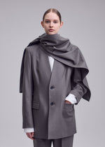 A person stands against a plain background wearing the MANNER grey oversized blazer and matching pants by ISSEY MIYAKE, crafted from upcycled fabric, plus a large grey scarf draped over their shoulders. Hair is pulled back; expression neutral.