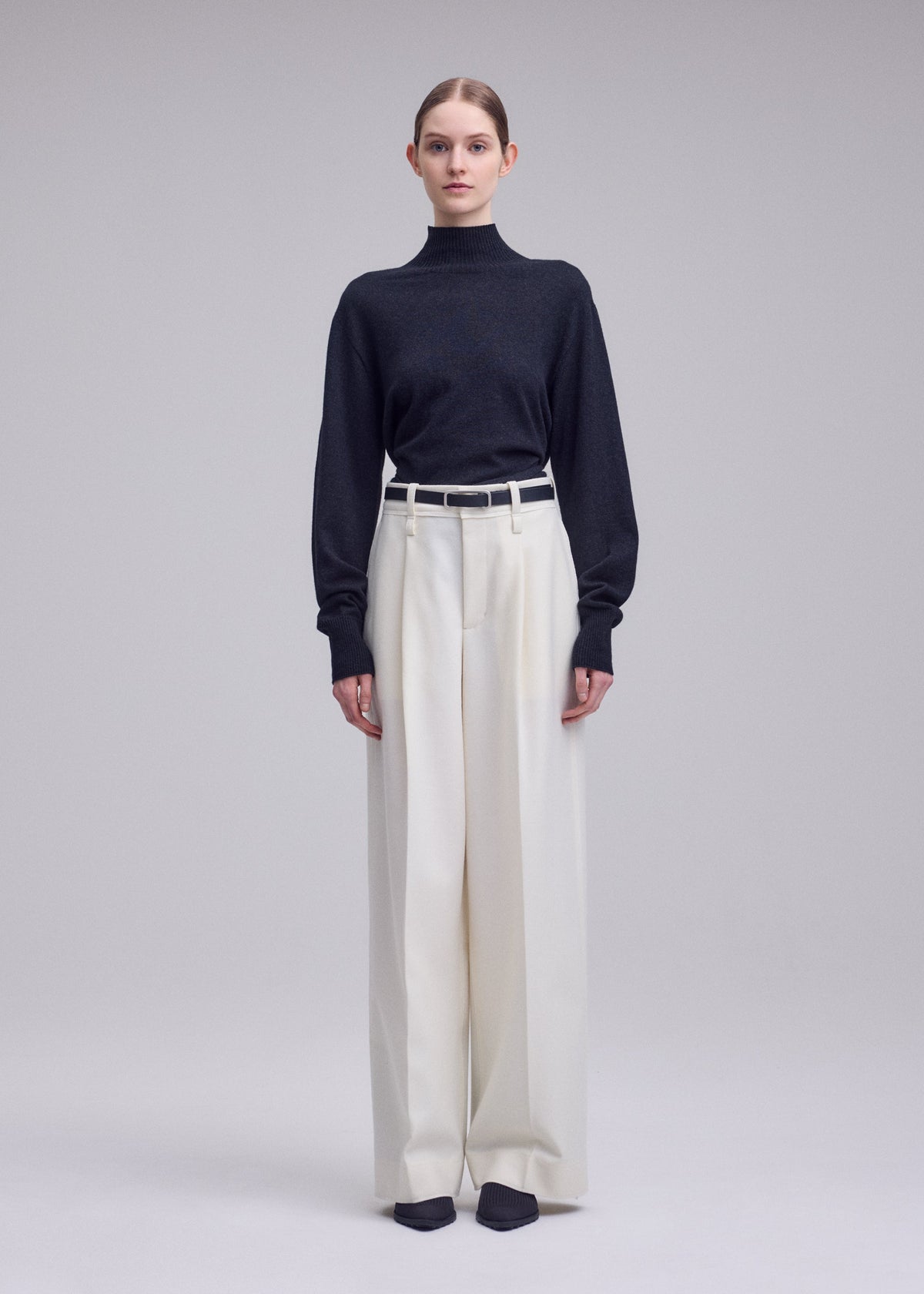A person stands facing forward against a plain light gray background, wearing the ISSEY MIYAKE SHELL black turtleneck sweater made of soft jersey material, tucked into high-waisted, wide-leg white straight pants with a white belt and black shoes. view 6 of 8