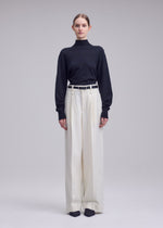 A person stands facing forward against a plain light gray background, wearing the ISSEY MIYAKE SHELL black turtleneck sweater made of soft jersey material, tucked into high-waisted, wide-leg white straight pants with a white belt and black shoes.