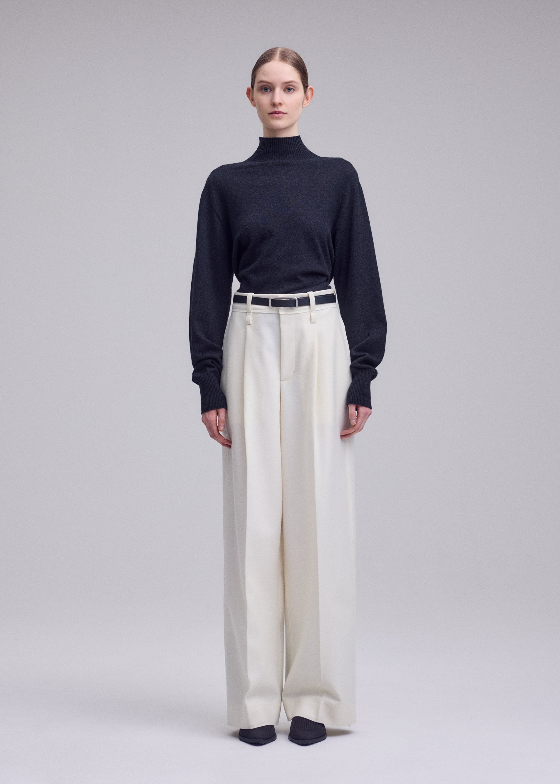 Zoomed view A person stands facing forward against a plain light gray background, wearing the ISSEY MIYAKE SHELL black turtleneck sweater made of soft jersey material, tucked into high-waisted, wide-leg white straight pants with a white belt and black shoes.