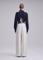 A person with long hair in a low ponytail stands facing away against a plain gray background, wearing the SHELL black top with a back cutout and white high-waisted, wide-leg trousers in soft jersey material by ISSEY MIYAKE.