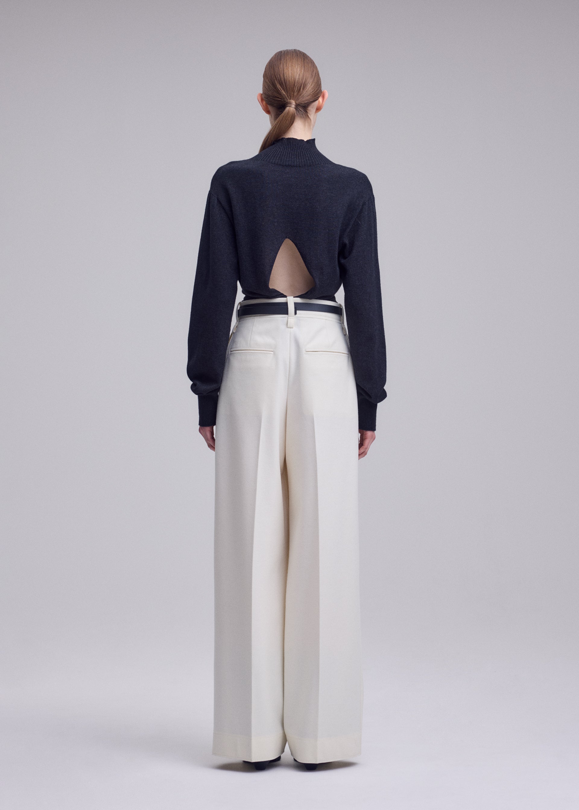 Zoomed view A person with long hair in a low ponytail stands facing away against a plain gray background, wearing the SHELL black top with a back cutout and white high-waisted, wide-leg trousers in soft jersey material by ISSEY MIYAKE.