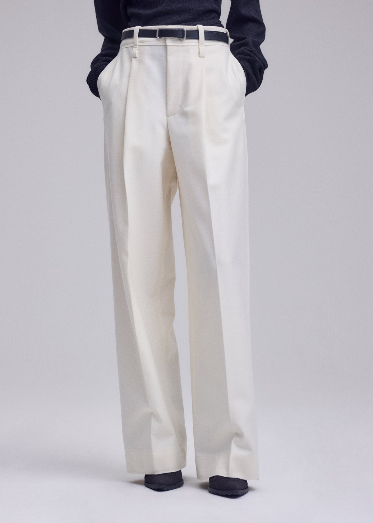 A person wearing the ISSEY MIYAKE SHELL high-waisted, straight white pants with hands tucked in the pockets, styled with a black belt and a dark long-sleeve jersey top. Only the lower body is visible. view 8 of 8