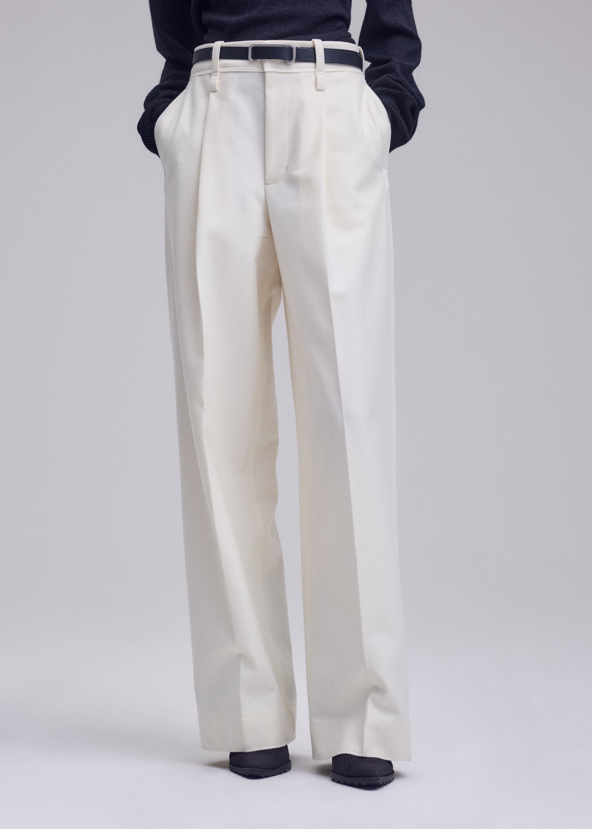 Zoomed view A person wearing the ISSEY MIYAKE SHELL high-waisted, straight white pants with hands tucked in the pockets, styled with a black belt and a dark long-sleeve jersey top. Only the lower body is visible.