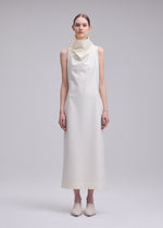 A person stands facing forward, dressed in the ISSEY MIYAKE SHELL—a sleeveless, ankle-length off-white dress made from soft jersey fabric, featuring a draped cowl neck—paired with matching shoes, set against a plain light grey background.