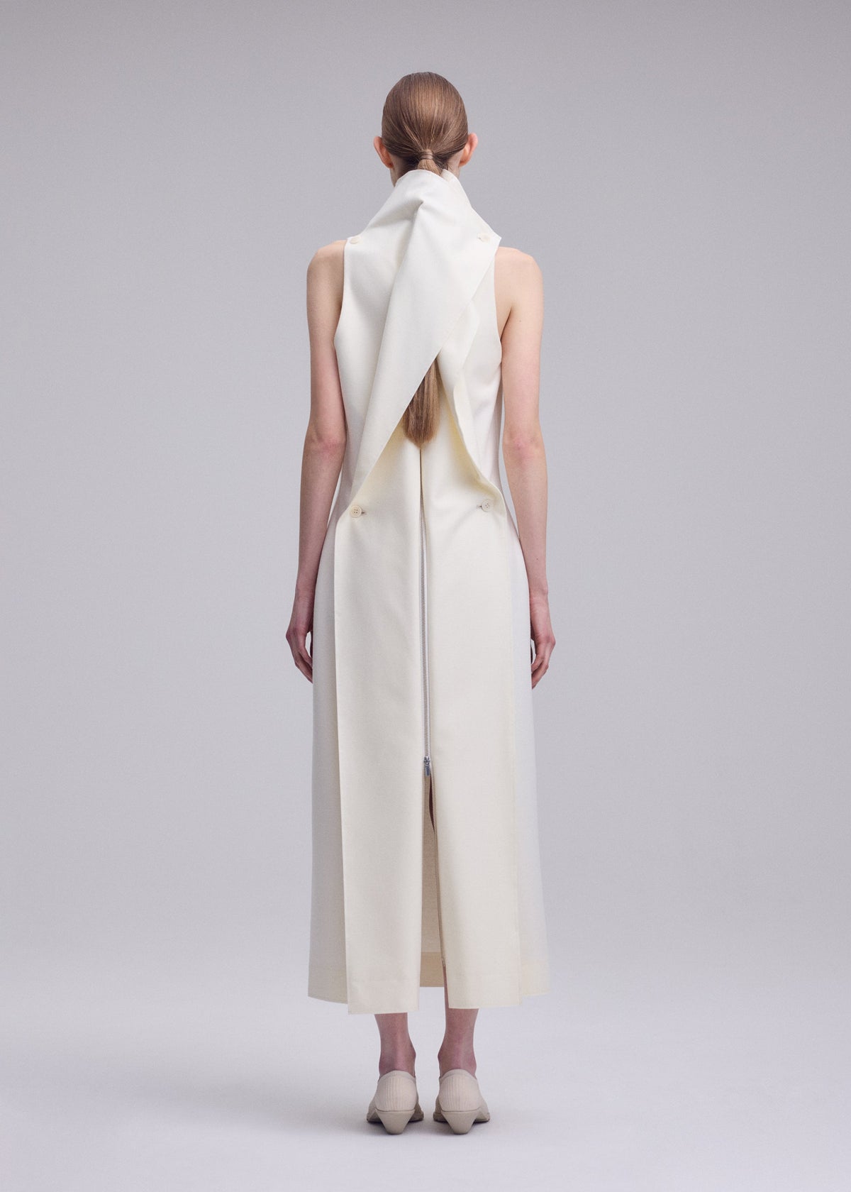 A person with long brown hair stands facing away, wearing the sleeveless, ankle-length white SHELL dress by ISSEY MIYAKE, featuring a sculptural collar and back detailing, paired with cream shoes against a plain gray background. view 6 of 7
