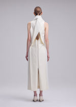 A person with long brown hair stands facing away, wearing the sleeveless, ankle-length white SHELL dress by ISSEY MIYAKE, featuring a sculptural collar and back detailing, paired with cream shoes against a plain gray background.