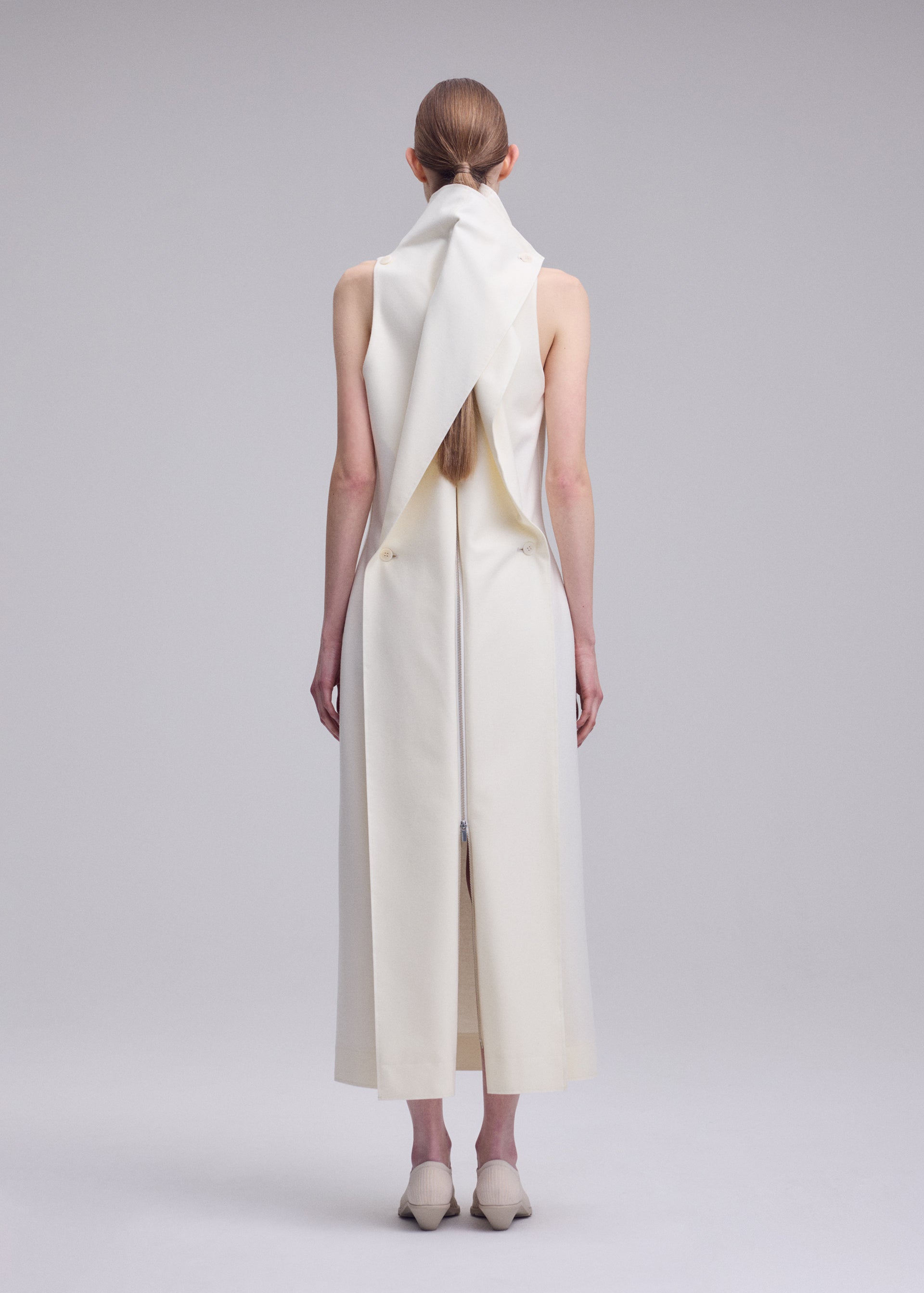 Zoomed view A person with long brown hair stands facing away, wearing the sleeveless, ankle-length white SHELL dress by ISSEY MIYAKE, featuring a sculptural collar and back detailing, paired with cream shoes against a plain gray background.