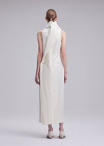 A person with tied-back hair models the SHELL dress by ISSEY MIYAKE—a long, sleeveless jersey piece featuring an asymmetrical draped collar and paired with white shoes. Facing away from the camera against a plain light background, they embody ISSEY MIYAKE’s signature minimalist elegance.