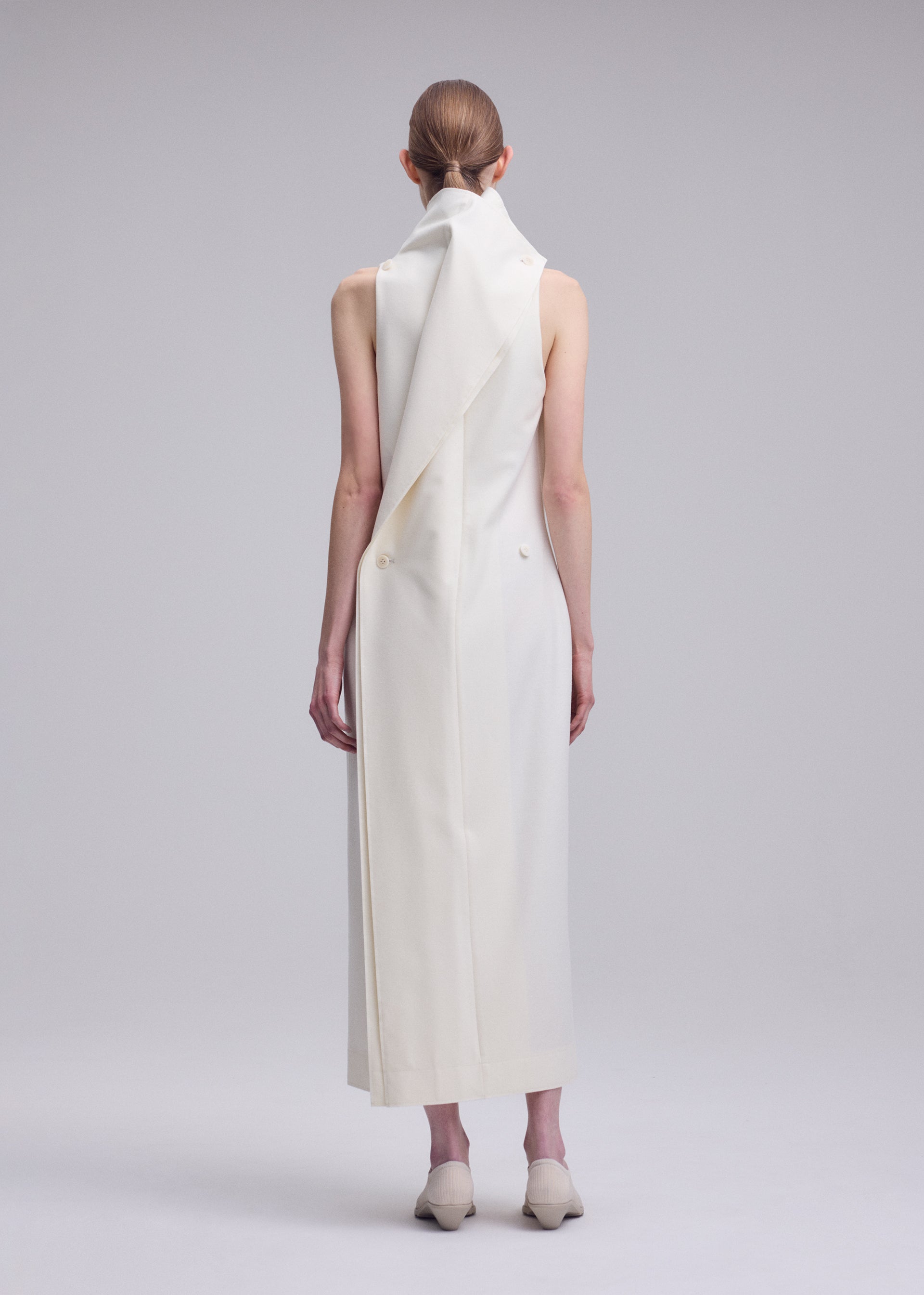 Zoomed view A person with tied-back hair models the SHELL dress by ISSEY MIYAKE—a long, sleeveless jersey piece featuring an asymmetrical draped collar and paired with white shoes. Facing away from the camera against a plain light background, they embody ISSEY MIYAKE’s signature minimalist elegance.