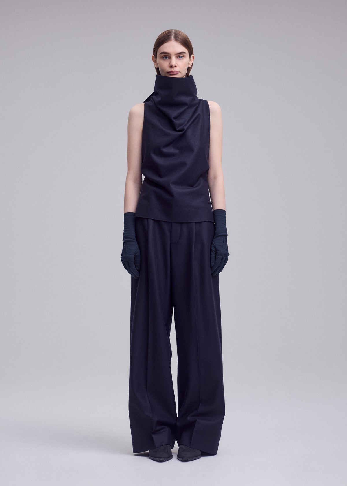 A woman stands against a plain background wearing SHELL by ISSEY MIYAKE: a sleeveless, high-neck black top made from soft jersey material, paired with wide-leg black trousers, black gloves, and pointed shoes. Her straight hair is parted down the middle, capturing the minimalist elegance of ISSEY MIYAKE. view 5 of 8