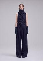 A woman stands against a plain background wearing SHELL by ISSEY MIYAKE: a sleeveless, high-neck black top made from soft jersey material, paired with wide-leg black trousers, black gloves, and pointed shoes. Her straight hair is parted down the middle, capturing the minimalist elegance of ISSEY MIYAKE.