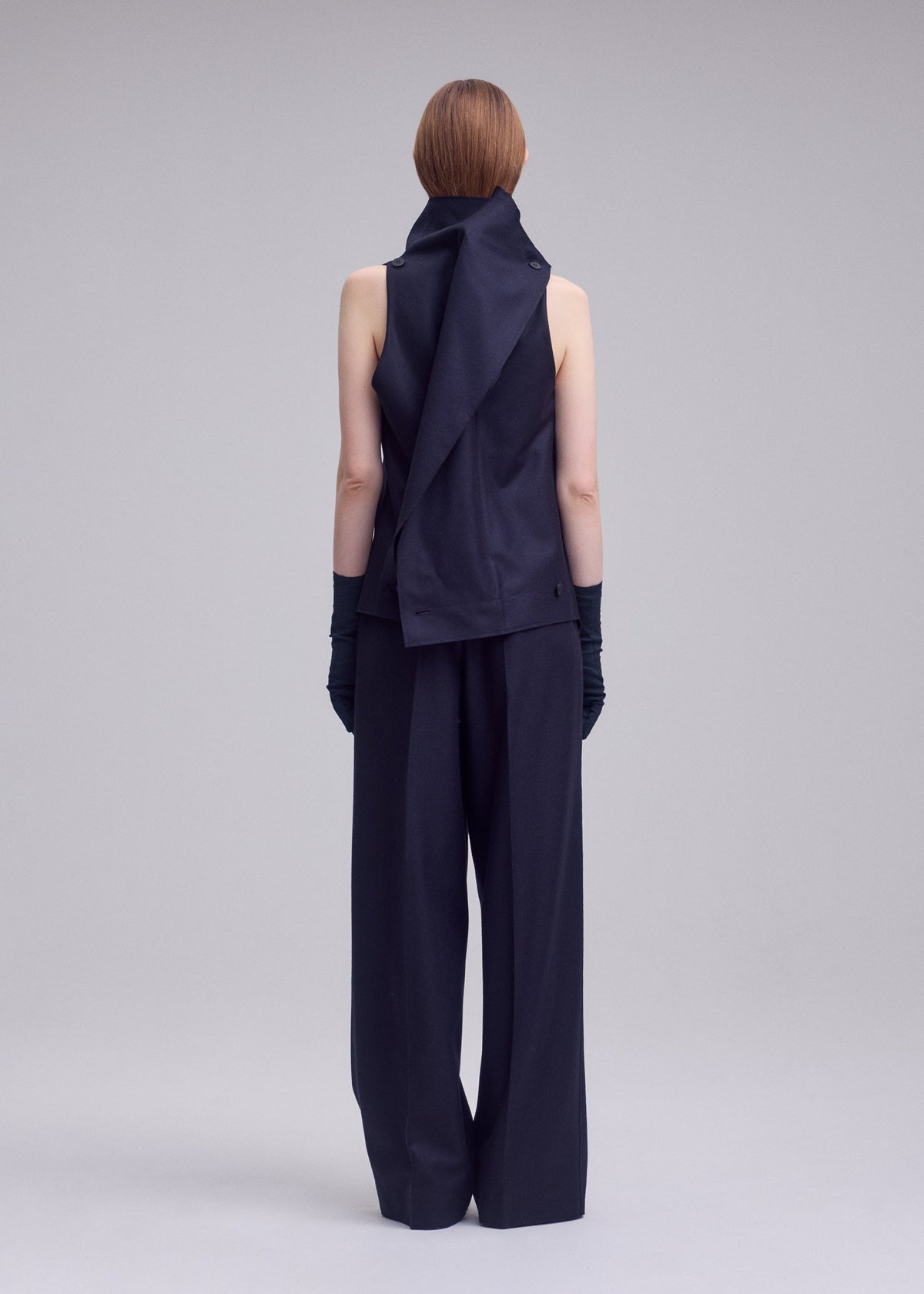 A person with short, straight, light brown hair stands facing away against a plain light gray background, wearing the ISSEY MIYAKE SHELL—a sleeveless, asymmetrical black top crafted from jersey material—paired with wide-leg black pants and black gloves. view 6 of 8