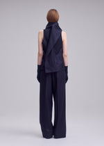 A person with short, straight, light brown hair stands facing away against a plain light gray background, wearing the ISSEY MIYAKE SHELL—a sleeveless, asymmetrical black top crafted from jersey material—paired with wide-leg black pants and black gloves.