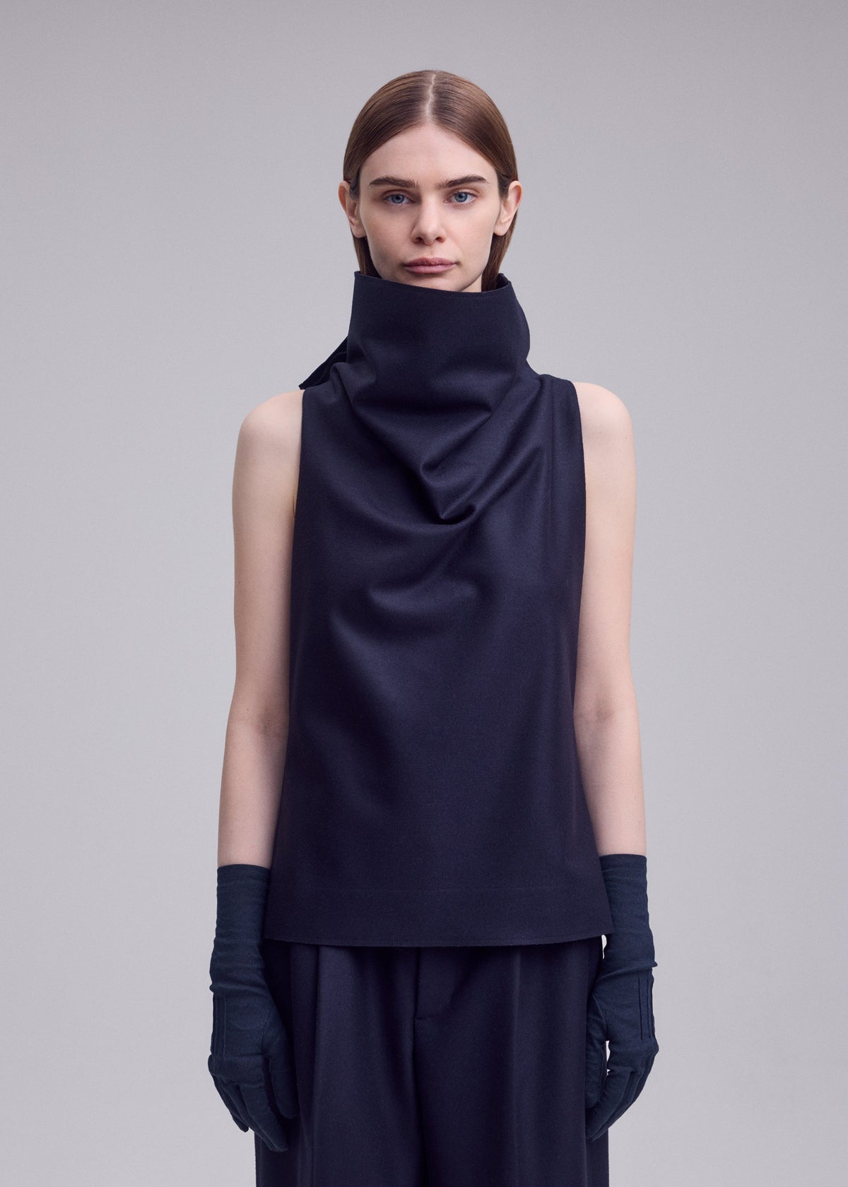 A person with straight brown hair wears the ISSEY MIYAKE SHELL sleeveless jersey top, paired with black pants and black gloves, standing against a plain light gray background in signature ISSEY MIYAKE style. view 7 of 8